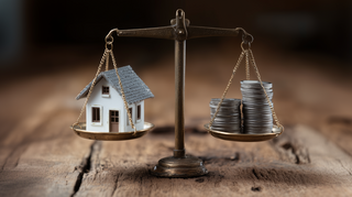 Eine antike bronzene Balkenwaage die in Balance steht. In der einen Waagschale ist ein kleines weisses Modell-Haus und in der anderen ein Stapel Münzen. Auf einem rustikalen Holztisch.